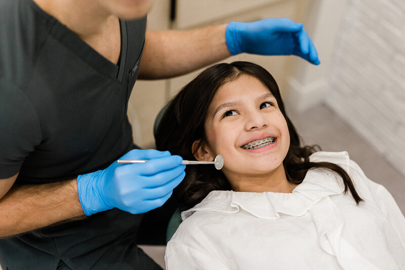 A young pediatric dentistry patient with braces smiles up at her Northside Chicago dentist, Dr. Michael Rabinowitz, DDS. The patient has long dark hair and wears a white ruffled blouse. Dr. Michael's gloved hands and inspection mirror frame the image.