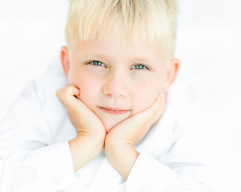 Boy with blonde hair and blue eyes wearing a white suit and lying on a white blanket outdoors.