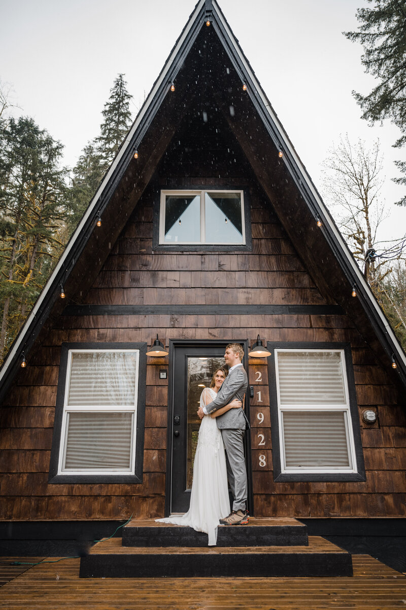 Bride and groom snuggled up in front of their adorable A-Frame cabin in Index, Washington during their rainy elopement day