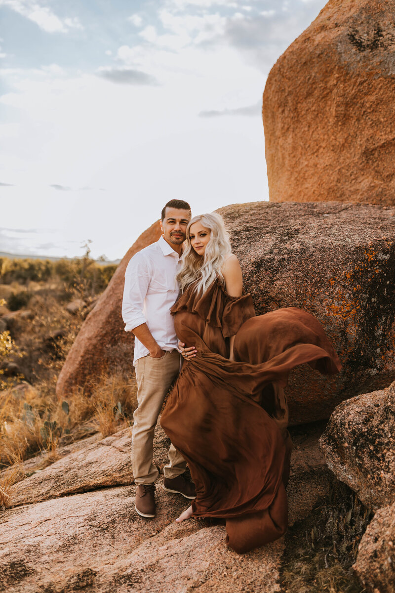 pregnant mother with a flowy dress blowing in the wind stands on a boulder with her husband