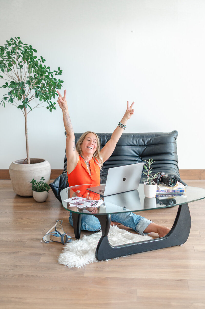 Woman in orange top sitting at coffee table on her laptop holding her hands in the air with peace signs.