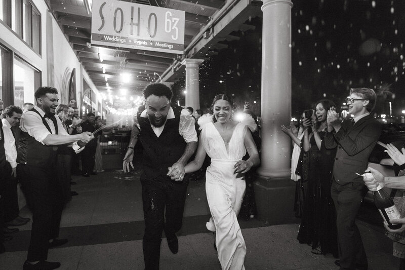 A newlywed couple runs hand-in-hand through a cheering crowd during their grand exit at SoHo63, a Chandler wedding and event venue. The black-and-white photo captures the joy and celebration under the venue’s glowing marquee, showcasing the magic of downtown Chandler weddings.