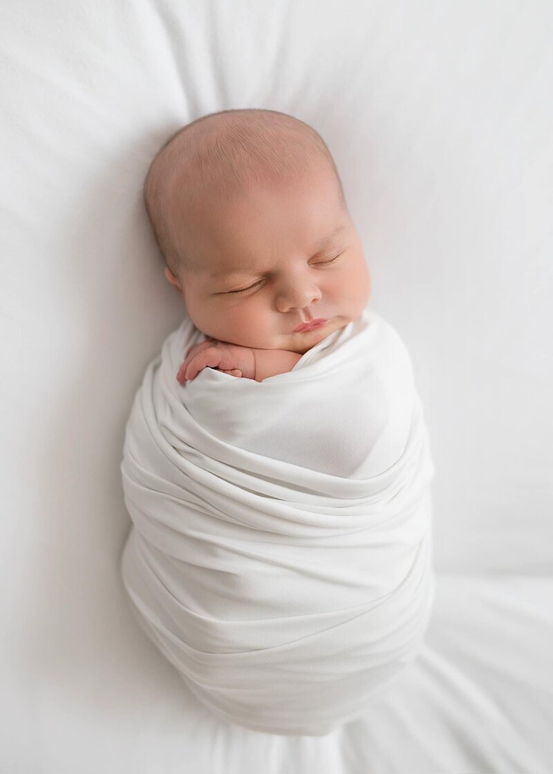 Baby boy wrapped in white swaddle sleeps on white beanbag.
