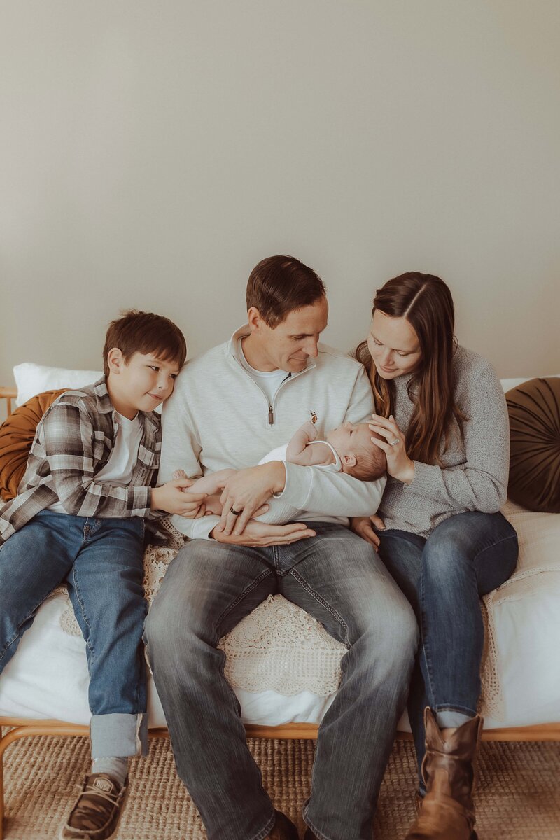 A family of four sits on a couch. The father holds a baby, with the mother and older child sitting beside him. The older child touches the baby's hand. Everyone appears calm and content.