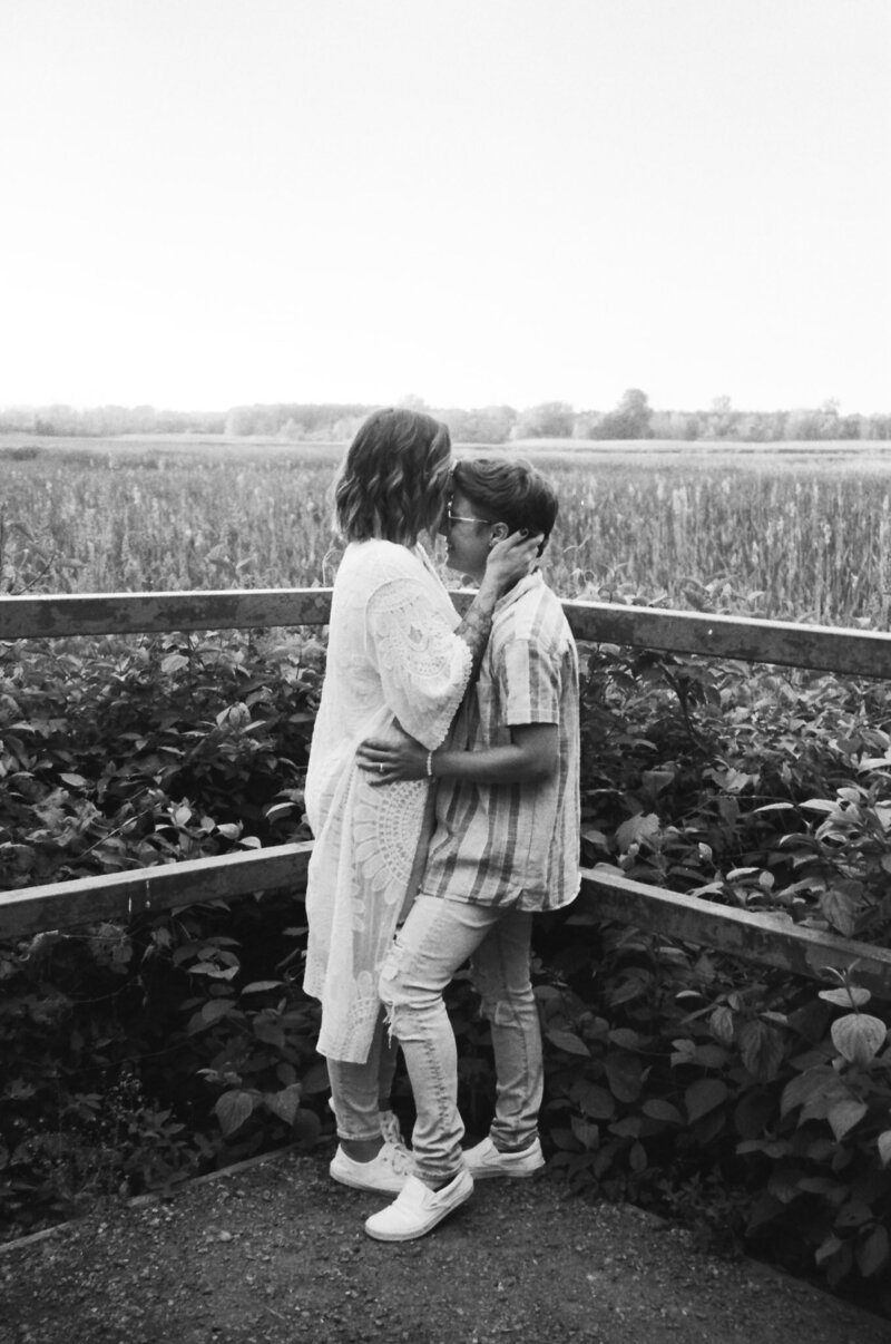 Two people stand closely by a wooden fence in a field, sharing an affectionate moment at Lynde Shores for an engagement photography session. One wears a long dress, the other a plaid shirt and jeans. Captured in black and white, this intimate scene was captured by Toronto engagement photographer, Jennifer van Son.