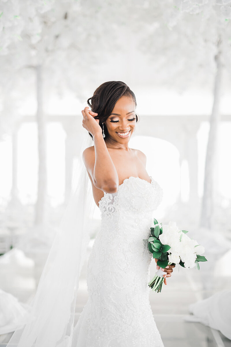 A bride looks down and touches her hair in an all white room.
