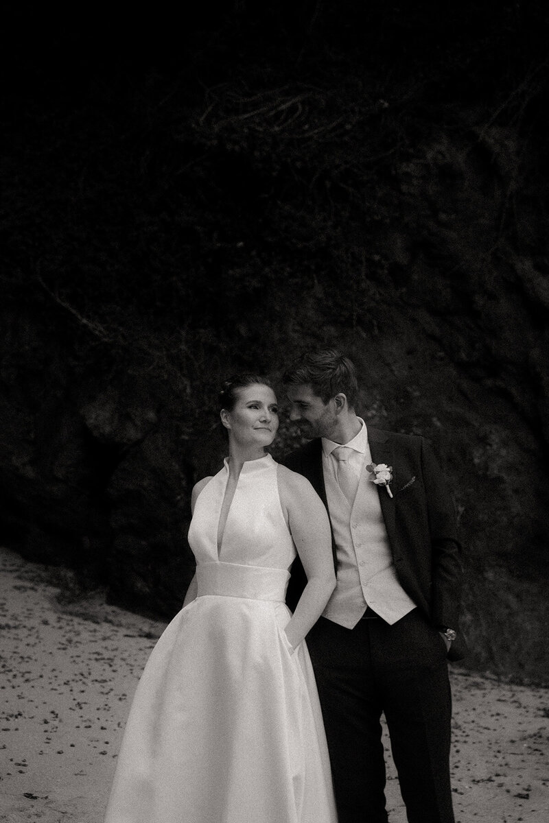 Clarisse et Corentin posant en tenue de mariés devant un rocher à la plage. Photographie de mariage vintage en noir et blanc, château de Beaussais, côtes d'Armor, Bretagne.