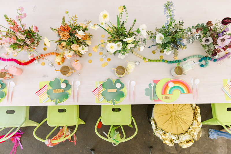 A St. Patrick’s Day-themed kids’ party table is adorned with clover-shaped plates, rainbow paper decor, and floral centerpieces arranged in a rainbow gradient. Personalized nameplates, gold coins, and disco balls add a playful touch to this bright and festive setup. Captured by Dani Lacey Photographs.