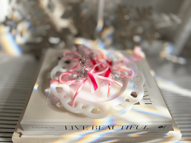 A decorative white plate with a pink peppermint candy cane bow holiday ornament from Sweet Pea Ink Creative sits on two stacked books, one titled "LIVE BEAUTIFUL," bathed in soft, natural light.
