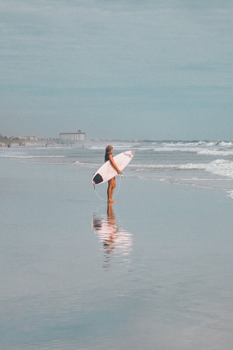 A surfer looks out to sea for waves while standing in a reflective area of the beach