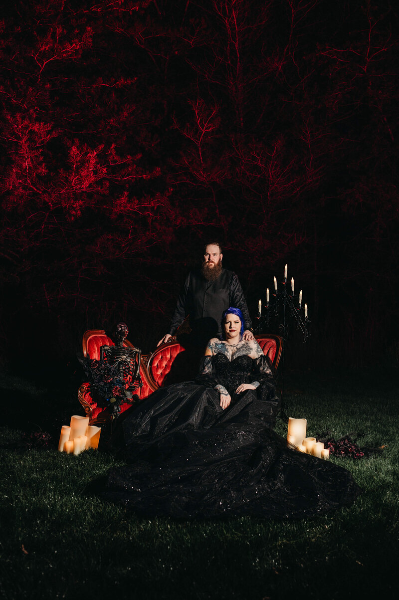 Bride in black dress sits son red couch for gothic style portrait at night.