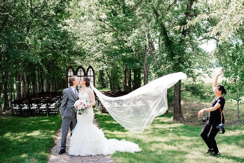 Behind the scenes photo of a photographer tossing a brides cathedral veil in the air for a dramatic photo at their outdoor ceremony location
