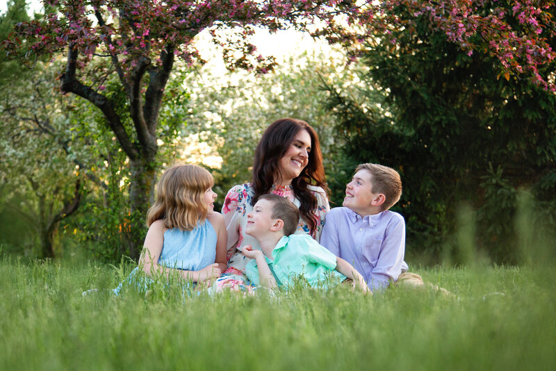 Mom and her three kids in pastels smiling at each other while sitting in grass with blossoming tree over them. 