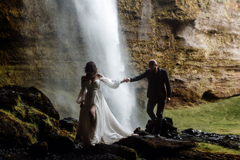 married couple is holding hands and being under a waterfall in Iceland