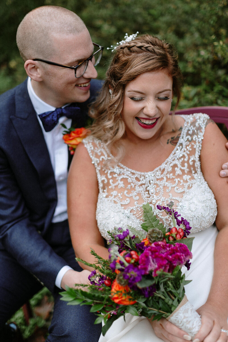 A bride in a beaded wedding gown holds a vibrant bouquet, while seated next to her partner in a formal suit, surrounded by greenery.