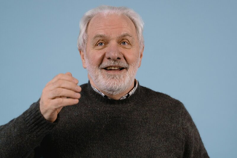 An older man with gray hair and a beard smiles and gestures while speaking against a blue background.
