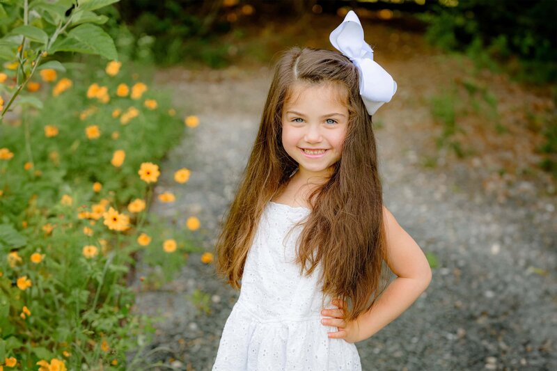 dark hair girl wearing white with a bow in her hair smiles with her hands on her hip in a flower garden