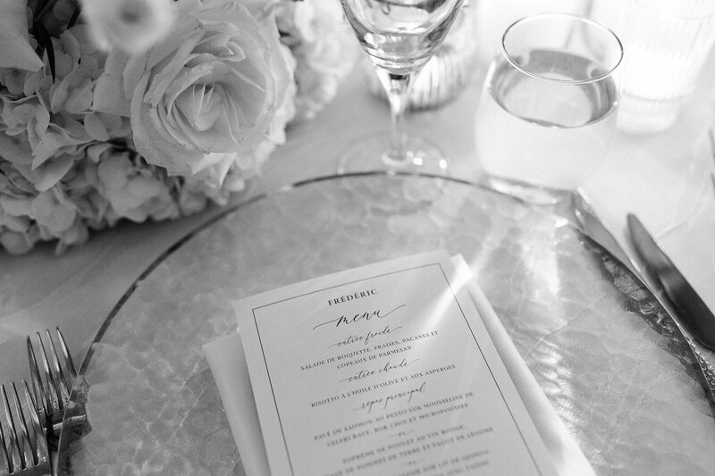 Elegant black and white photo of a wedding menu with soft floral arrangements and candles in the background. 