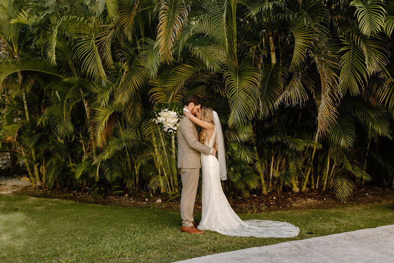 Bride and groom embrace in front of lush palm trees at their Cancun destination wedding.