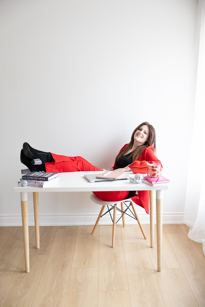 Emily is wearing a fully red suit and black boots with sparkly heels. Her legs and feet are propped up on the desk and she is leaning back in the white chair. There are books, disco balls, electronics, a pink lip pattern mug and red lip shaped phone on the desk in front of her.