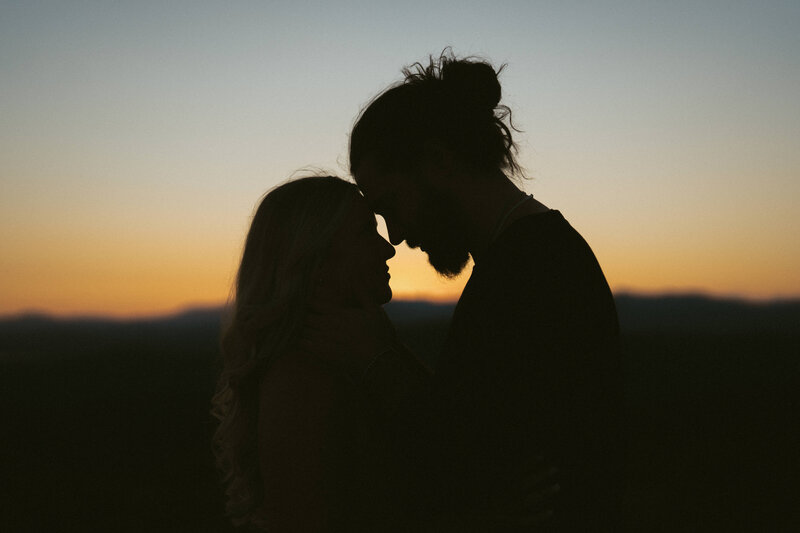 Silhouette of a couple with their foreheads together at sunset, with a warm orange glow behind them.