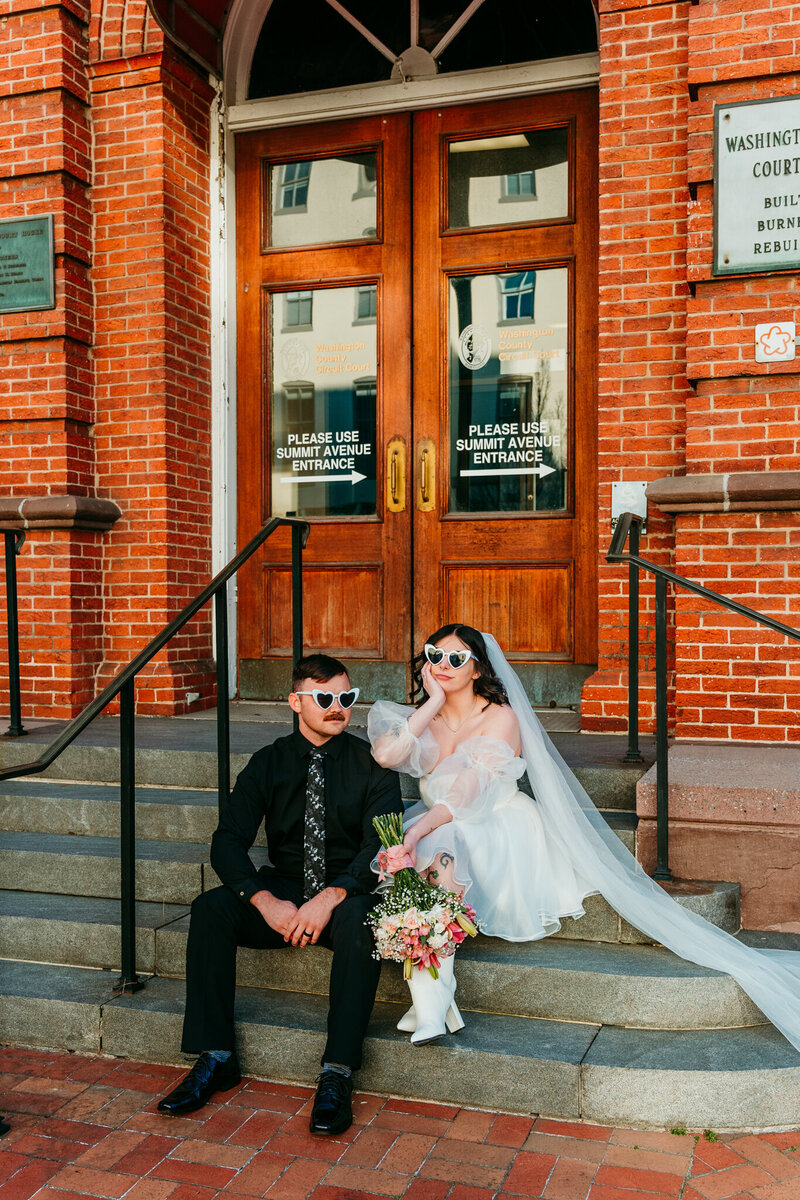 Stylish elopement couple sitting on courthouse steps with sunglasses and bouquet in downtown Hagerstown, Maryland