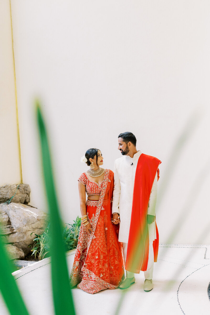 A couple dressed in traditional Indian wedding attire stand together outdoors. The bride wears a red lehenga and the groom wears a white sherwani with a red scarf. They hold hands while surrounded by greenery.