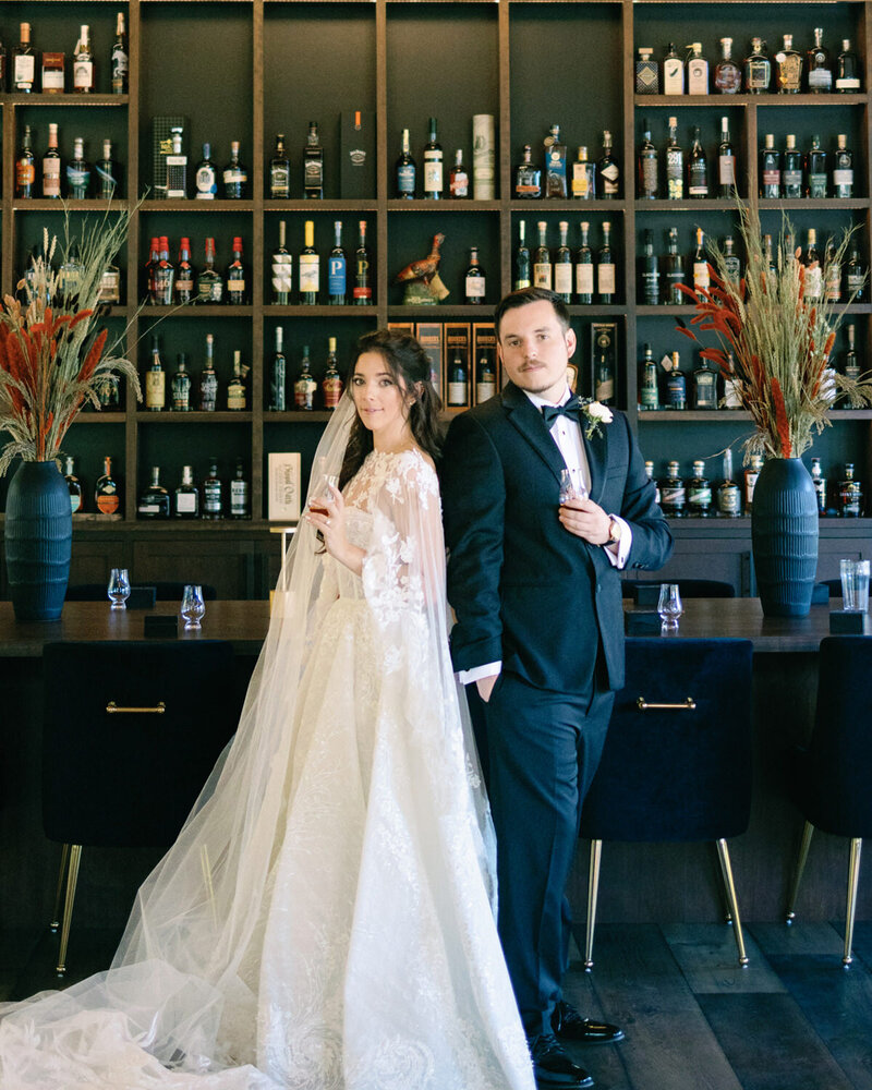 Couple is posing after their wedding in a bar area with bottles of wine and other mixtures in the background.