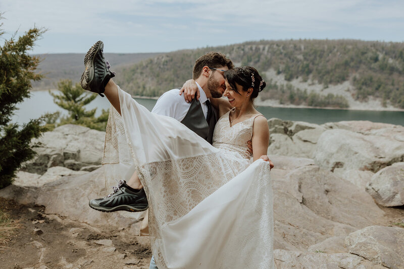 groom is lifting bride and kissing her cheek. Bride is wearing hiking boots and kicking out her feet.