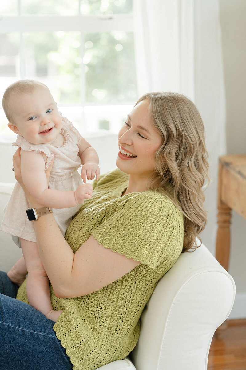 Mother holding her smiling baby girl up in the air while laying on a white couch