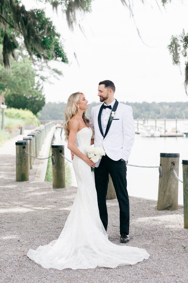 A beautiful bride in a fitted lace gown and a groom in a white tuxedo jacket share a loving moment by the waterfront at Spring Island, South Carolina. A timeless and sophisticated wedding captured by Amia Marcell Photography.