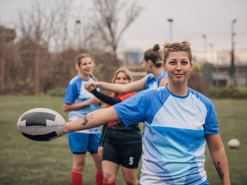 A female athlete holding an American football with three other athletes in the background.