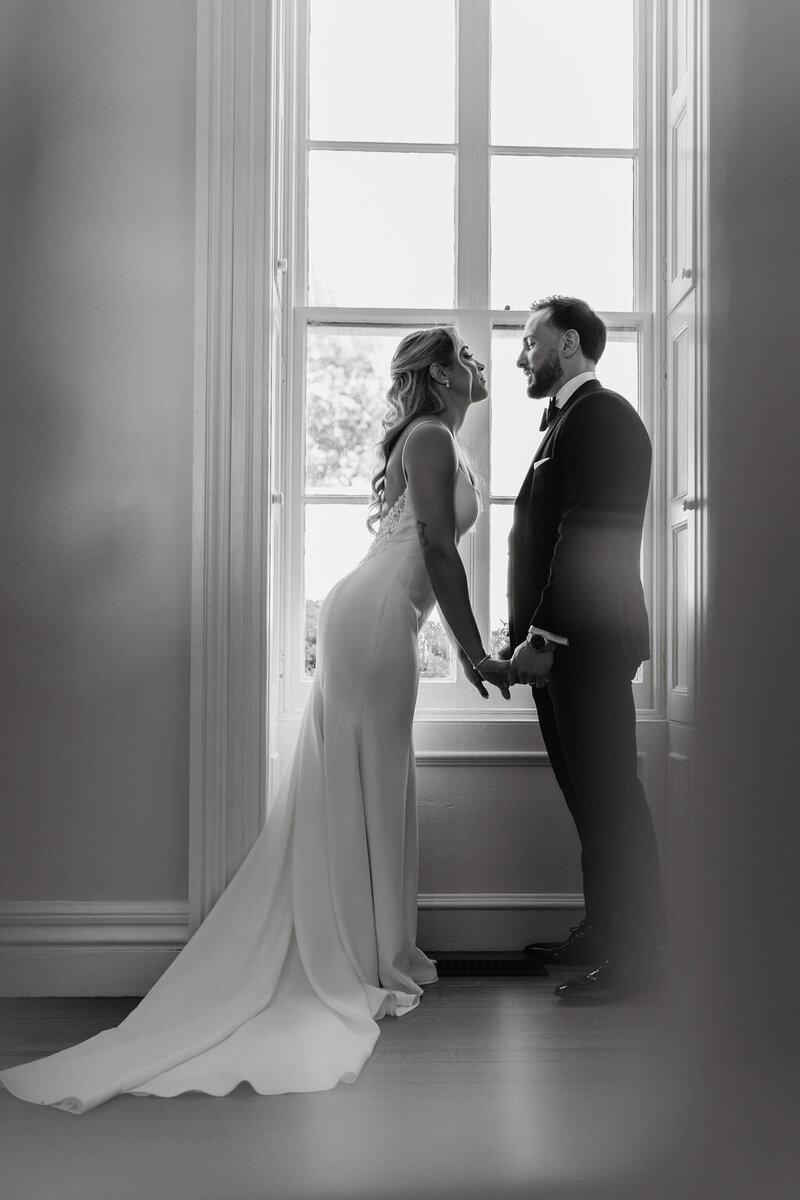 Black and white photo of newlywed couple leaning in for a kiss in front of a window in Long Island, NY | Emma LeeAnn Photography