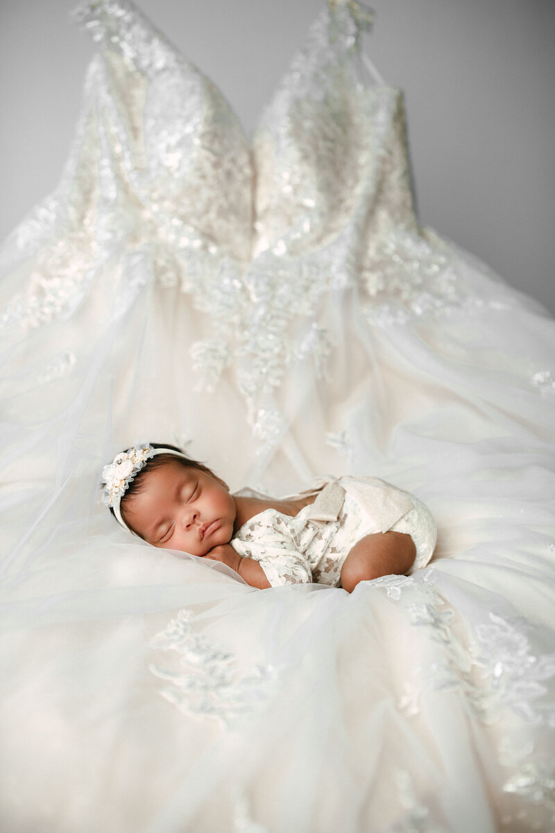 newborn baby girl in white laying in pose on top of mom's wedding dress  baby wearing lace onesie with lace bow laying on her tummy