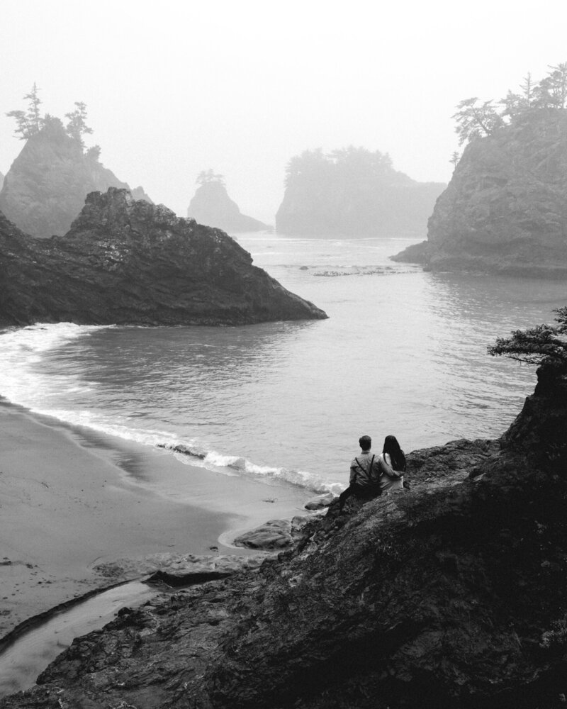 A couple sits on a large seastack and gazes out over a moody, rocky beach for their Oregon coast elopement.