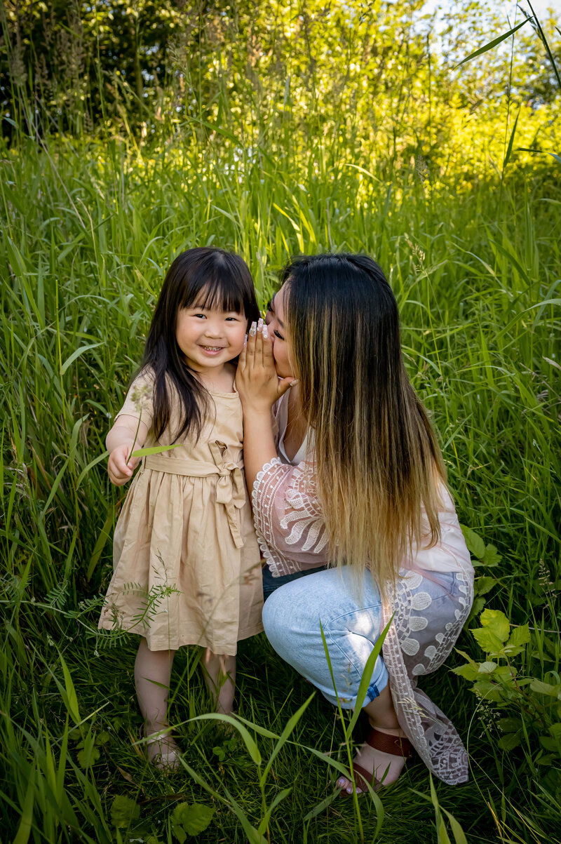 Mom telling her daughter a secret in her ear, they are in tall long green grass at Blackie Spit in Surrey, BC.