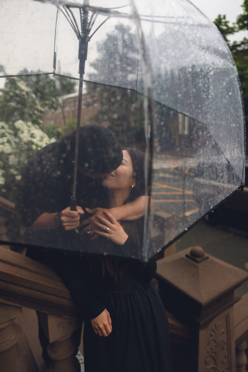 Engaged couple kissing under a clear umbrella during rainy day in Philadelphia