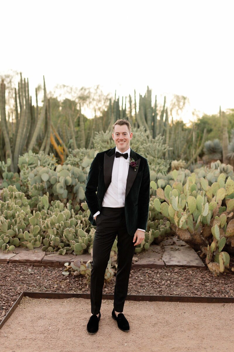 Groom stands with one hand in pocket in cactus garden.