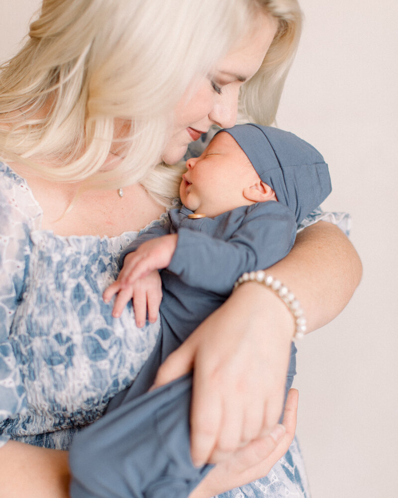 Closeup of mom in blue and white floral gown cradling her newborn baby in a blue Lou Lou and Co knotted onesie and nuzzling his head, by Newburyport family photographer Fieldstone Studio.
