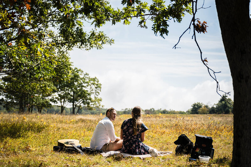 A couple on their elopement day sit on a park bench on top of a hill with their hiking backpacks. The couple wrap their arms around one another and face the view of the Blue Ridge Mountains from Heritage Park in Blacksburg, Virginia.