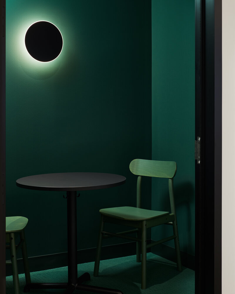 A small phone room featuring a wood table and chairs, soft lighting from a wall sconce, and forest green walls and carpet