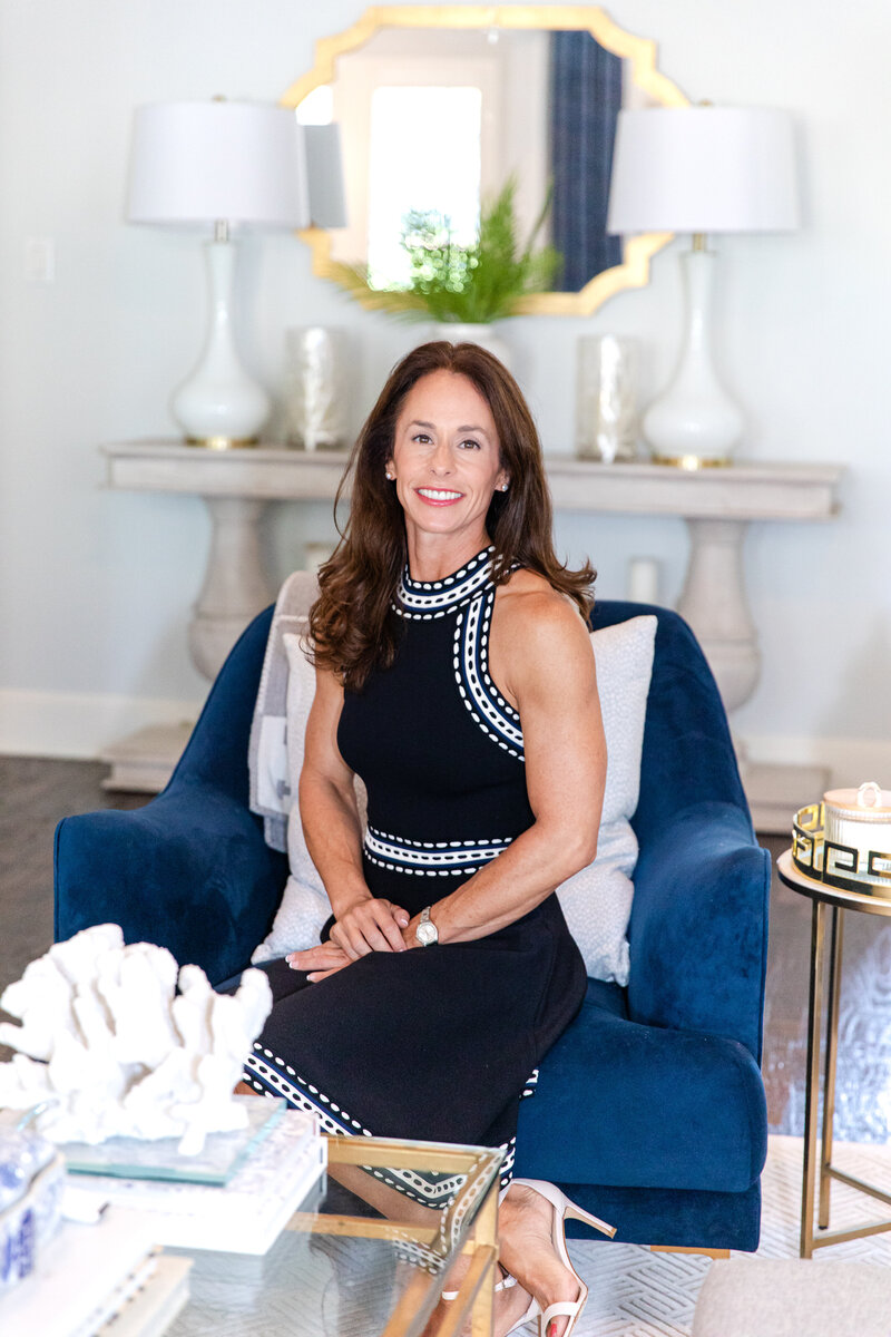 female realtor sitting on a blue chair wearing a black sleeveless dress looking at the camera smiling