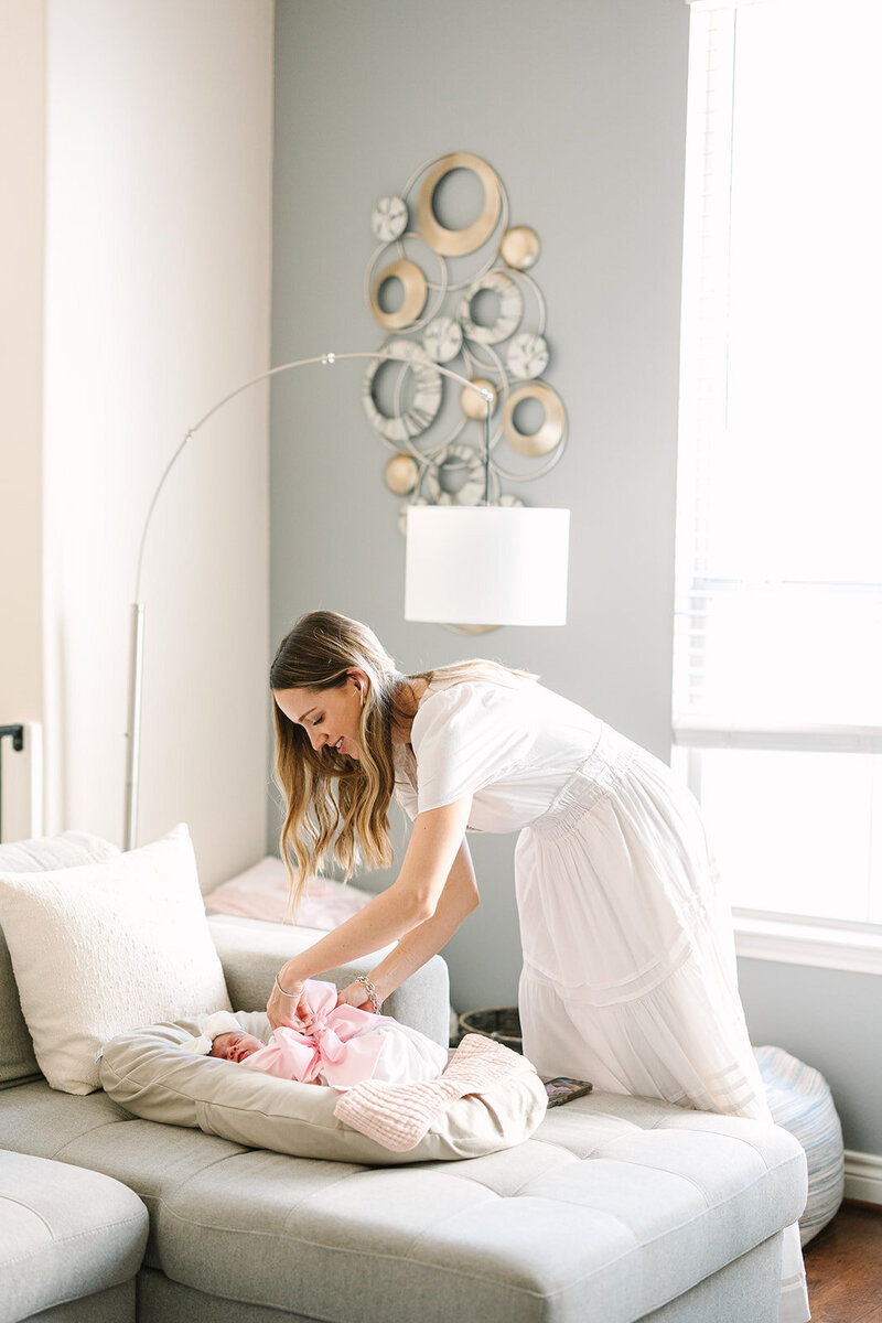 Mom in long white dress adjusting big pink bow on newborn baby laying on a baby pillow