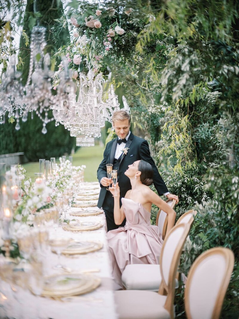 A couple at their table during their wedding at Villa Cetinale in Tuscany. Bride in a pink wedding dress is sitting and cheering with champagne with the groom. Beauty perfectly captured by luxury international wedding photographer Andreas K. Georgiou.