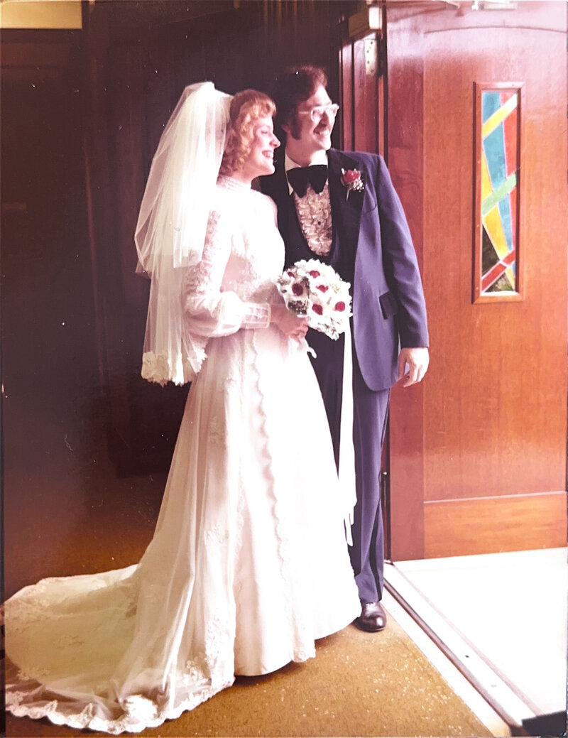 vintage photo of a bride and groom smiling at each other, the bride in a long-sleeved lace gown with a bouquet and veil, and the groom in a dark suit with a ruffled shirt and flower boutonniere