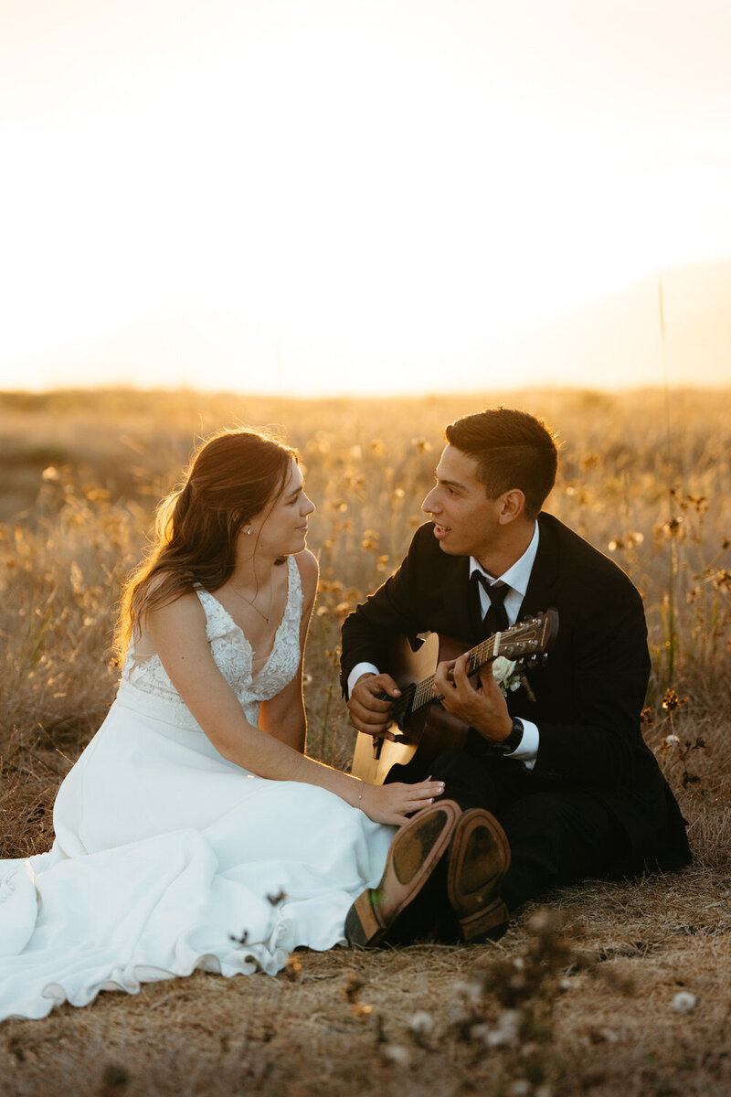 An Artistic Elopement Idea, groom plays his guitar for bride while sitting in a meadow, the golden grass behind them is lit up by the sunset