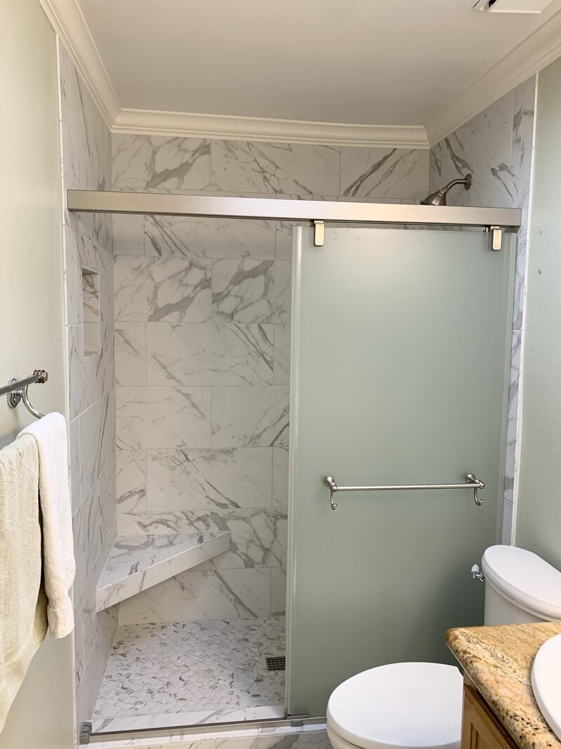 A faux marble shower with frosted glass sliding doors and brushed nickel hardware. The sliding door has a towel bar and there is a seat on the left hand corner of the shower made from faux marble.