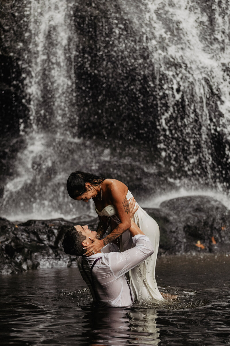 Wedding couple lifting bride out of the water in front of a waterfall