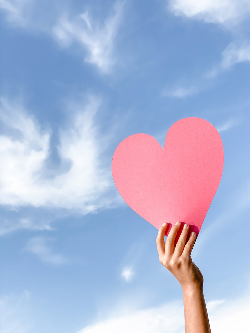Hand holding a heart cutout against blue skies and clouds, symbolizing heart-led insight, healing, and openness through the Stellar Insights podcast
