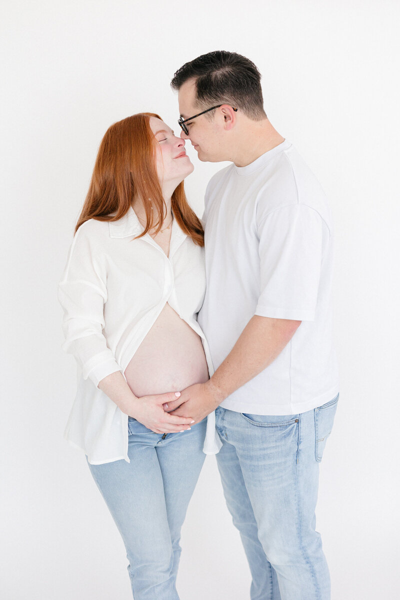 Expecting parents wearing white shirts and jeans on a white backdrop during their louisville maternity photography session